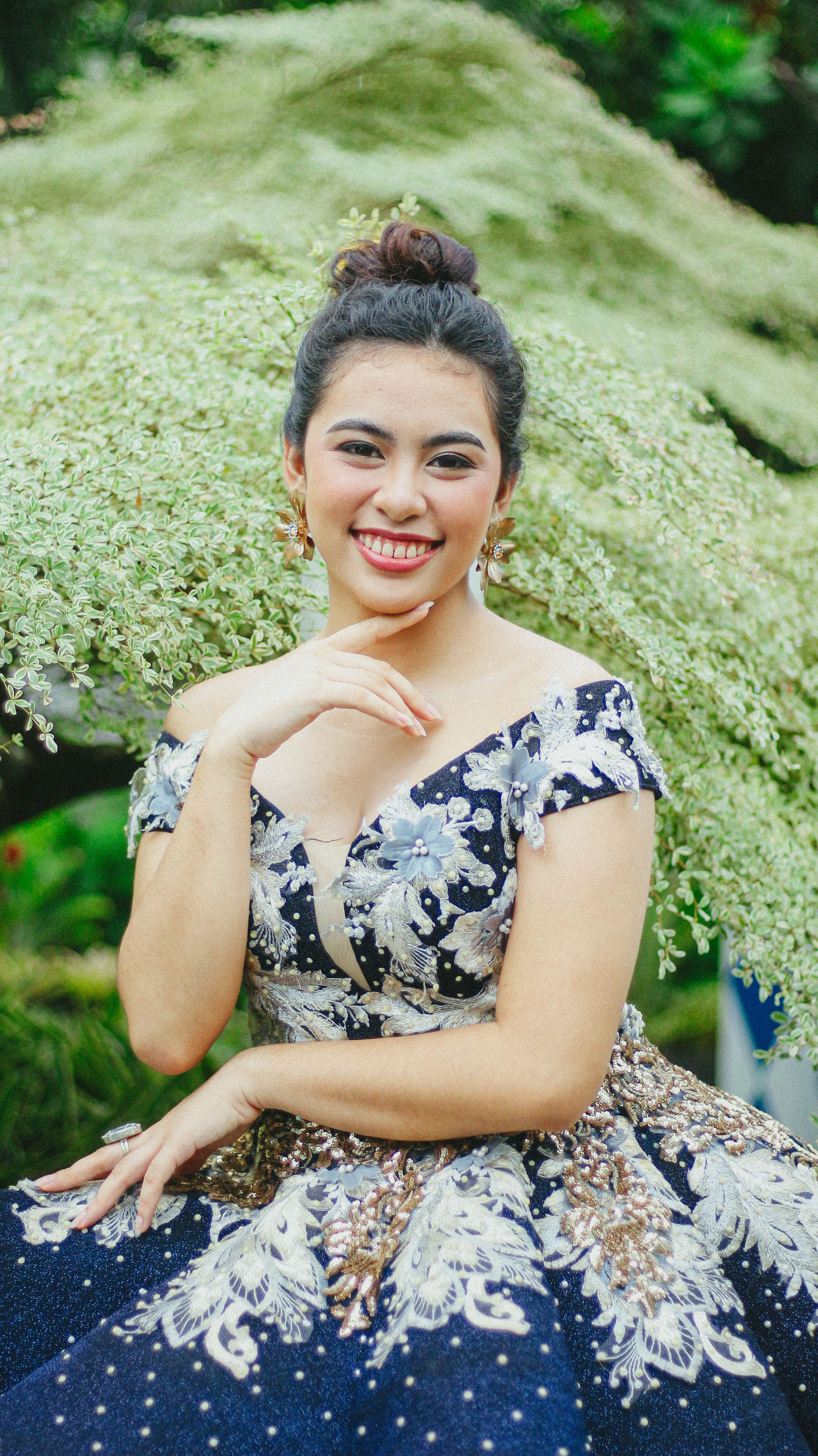 Smiling woman in a floral embroidered gown sitting outdoors amidst greenery.