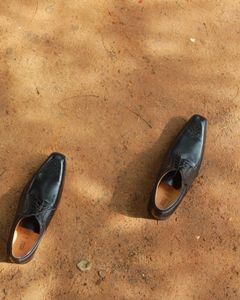 A pair of black leather shoes laid on an earthy surface under sunlight.