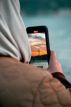 Person photographing a sunset over a bridge with a smartphone, highlighting modern connectivity.