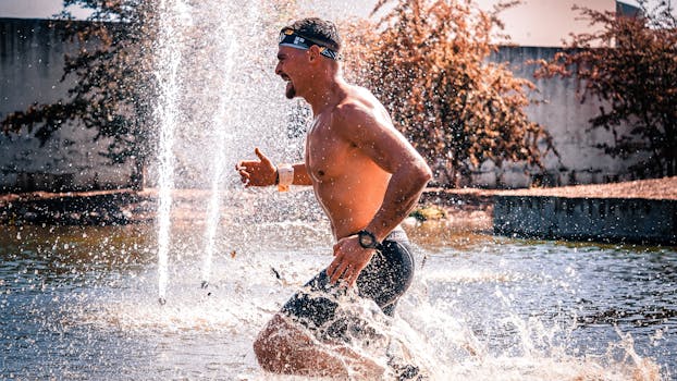 A shirtless man enjoying an energetic run through a water fountain outdoors.