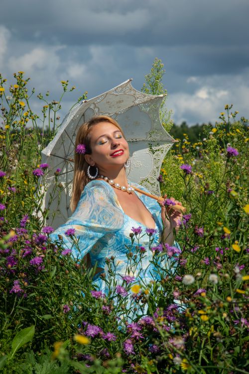 Free Serene woman in blue dress with lace parasol enjoying a sunny day in a flower-filled field. Stock Photo