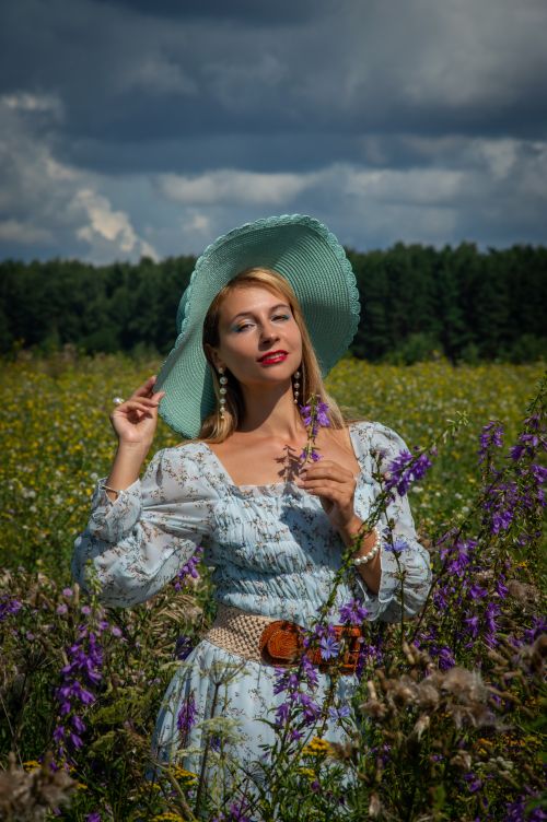 Free Elegant woman in blue floral dress and hat posing in a colorful meadow under a cloudy sky. Stock Photo