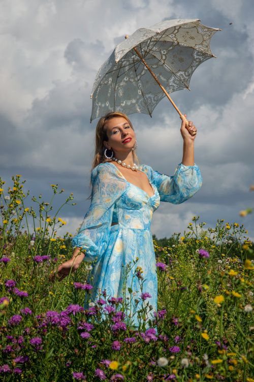 Free Woman in a blue floral dress holding a lace umbrella amidst wildflowers in daylight. Stock Photo