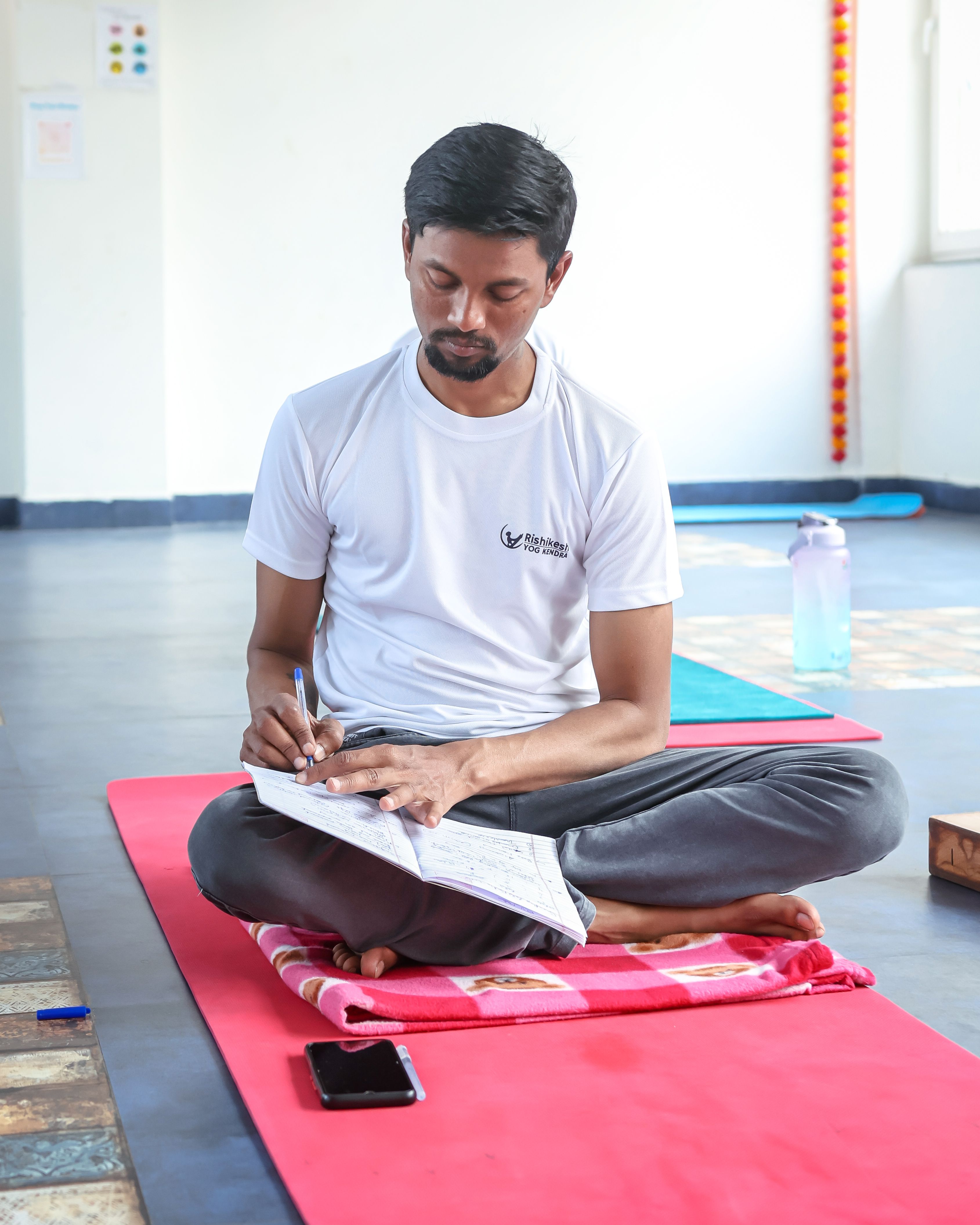 A man sitting cross-legged on a yoga mat, writing in a journal indoors.