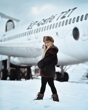 A young woman in fur clothing poses before a snow-covered airplane, showcasing winter fashion.