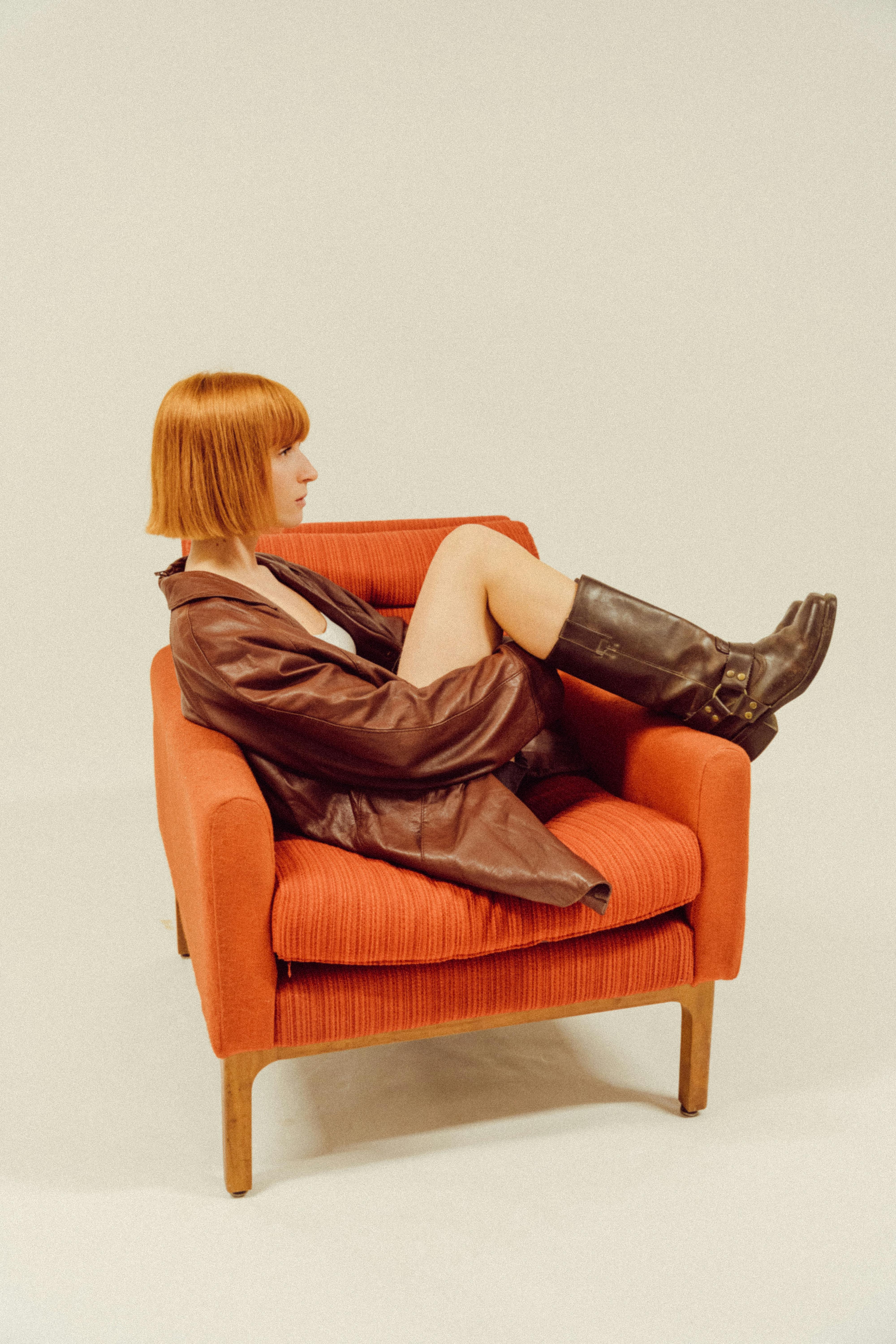 Trendy woman in leather jacket sitting comfortably on an orange chair indoors.