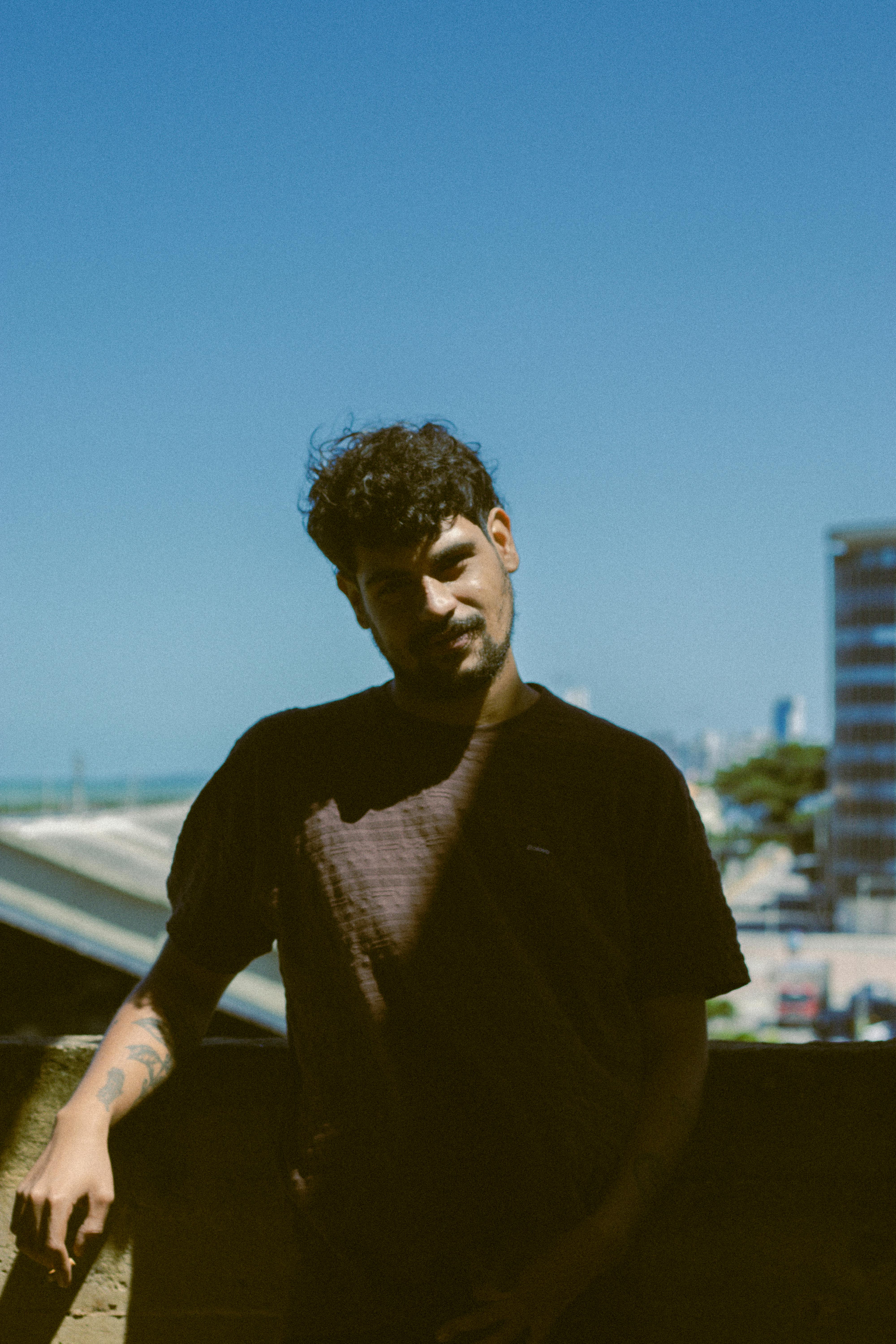 Casual portrait of a young man standing on a balcony with urban cityscape in background.