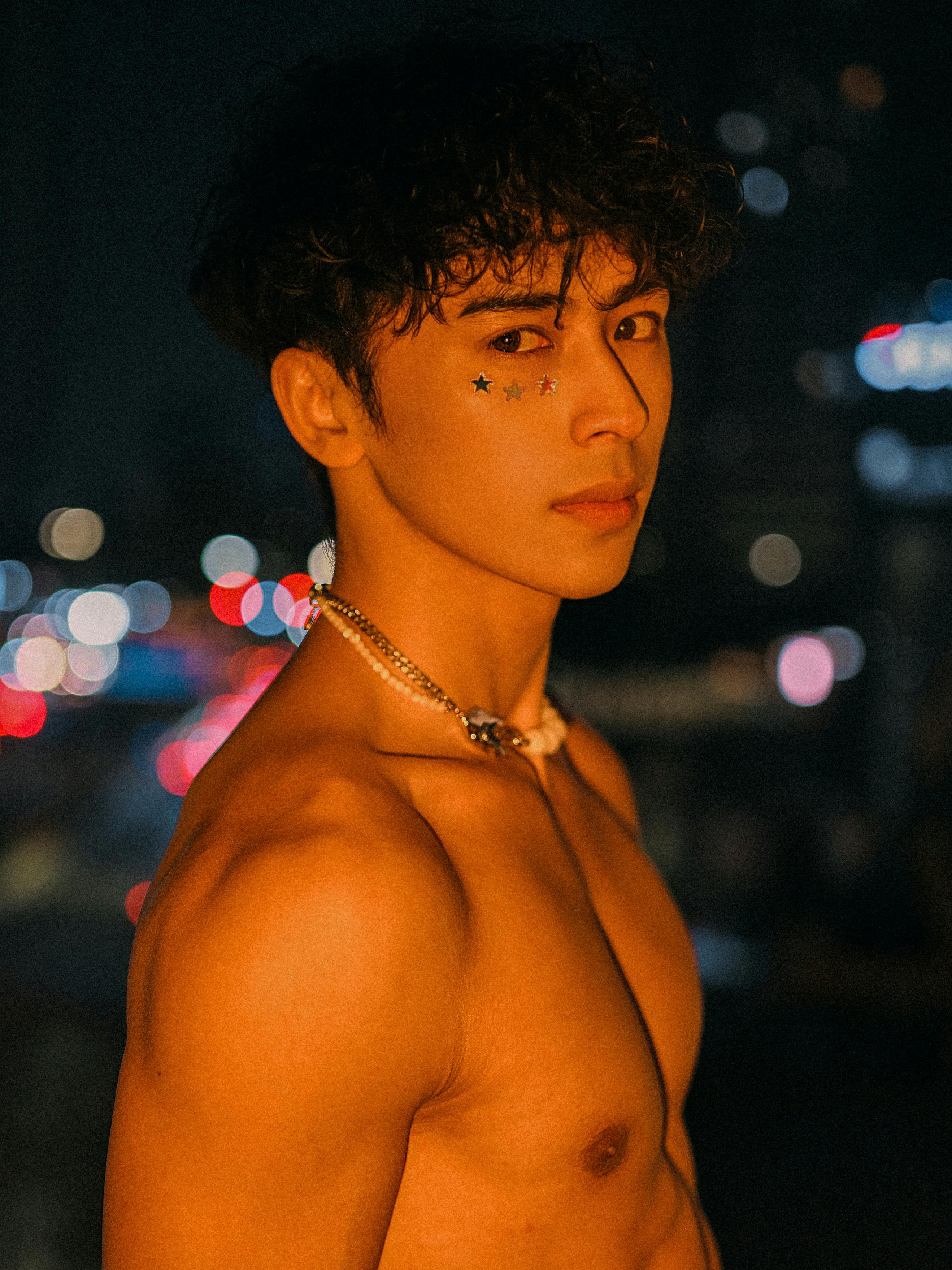 A young adult male poses under city lights with star stickers on face, creating a moody portrait.