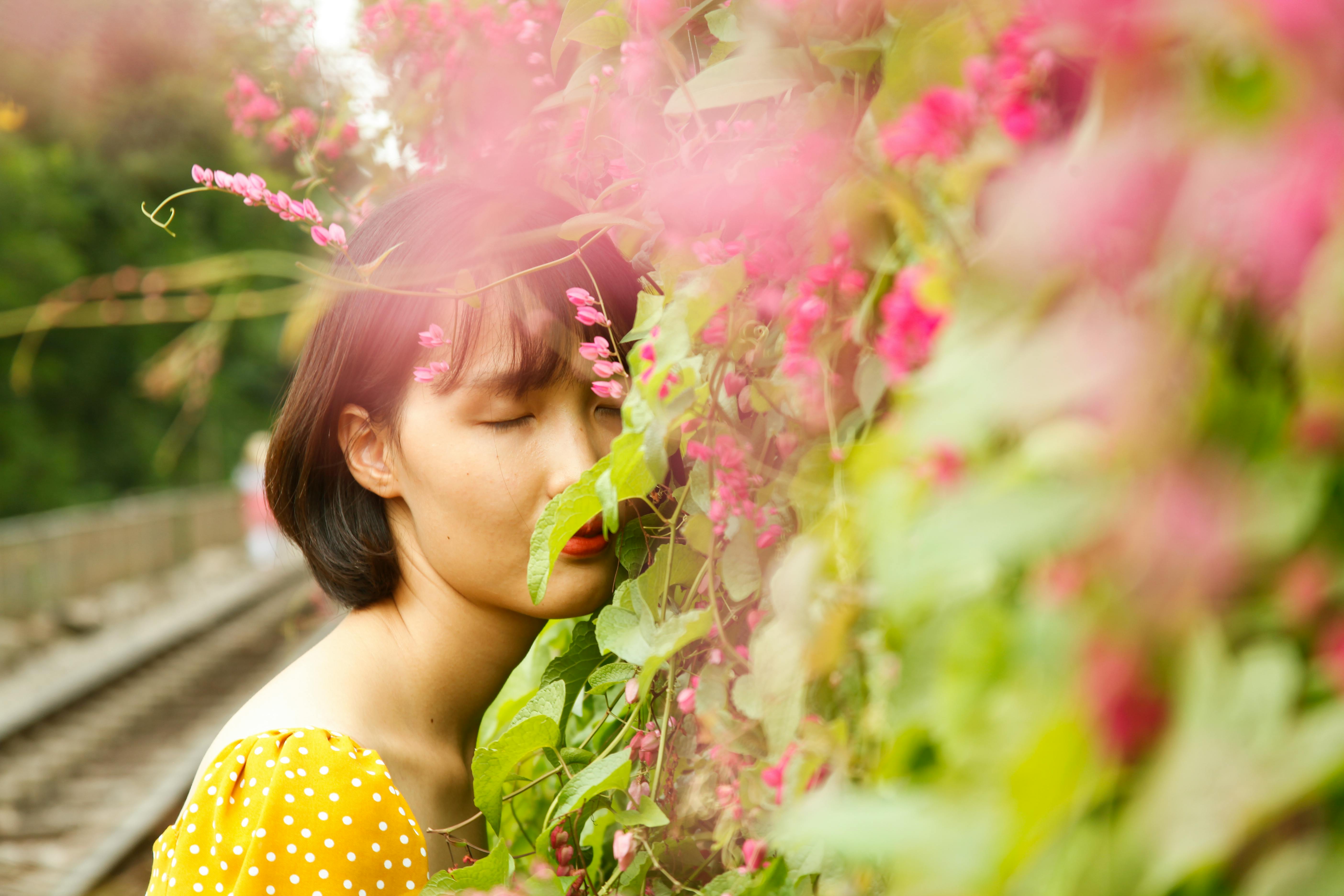 Asian woman in a yellow polka dot dress surrounded by blooming pink flowers outdoors.