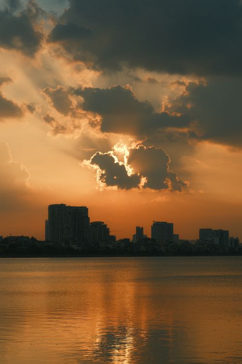 Free Dramatic sunset with sun rays breaking through clouds over a city skyline reflecting on calm water. Stock Photo