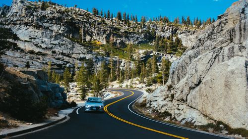 Free A car drives through a winding road in a mountainous landscape, surrounded by trees and rocks under a clear summer sky. Stock Photo