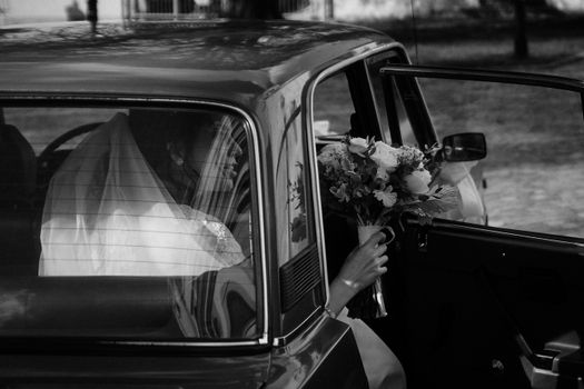 A bride with a bouquet sits in a vintage car, captured in a timeless black and white photograph.