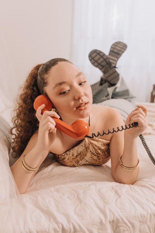 Free A woman lying on a bed talking on a vintage red telephone, wearing casual clothes indoors. Stock Photo