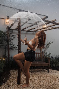 A vibrant young woman enjoying the rain under an umbrella in a garden setting.