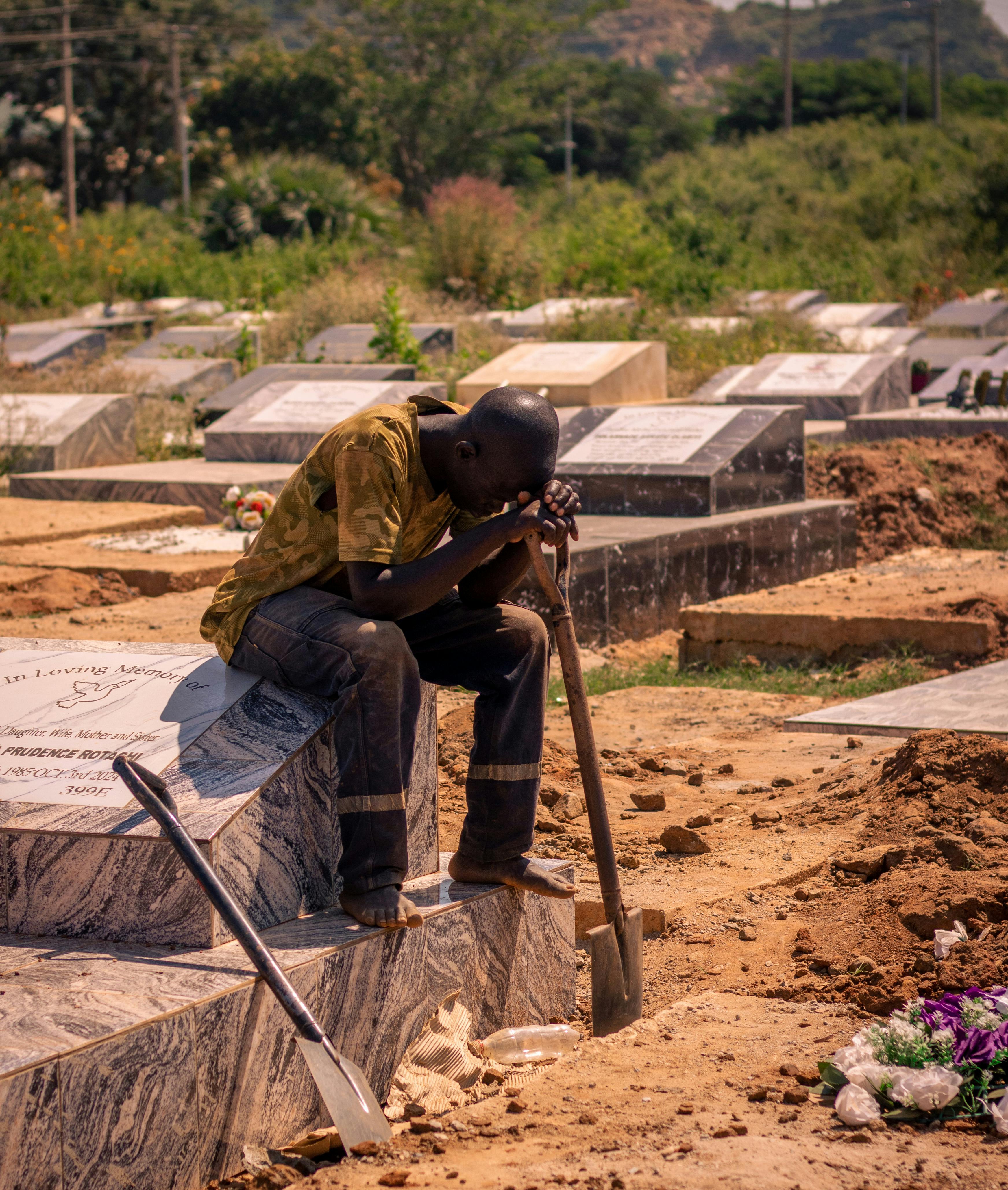 A man sits on a grave, lost in thought amidst a quiet cemetery landscape.