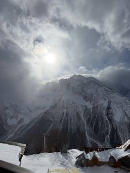A breathtaking view of snowy mountains under a cloudy sky with the sun peeking through.