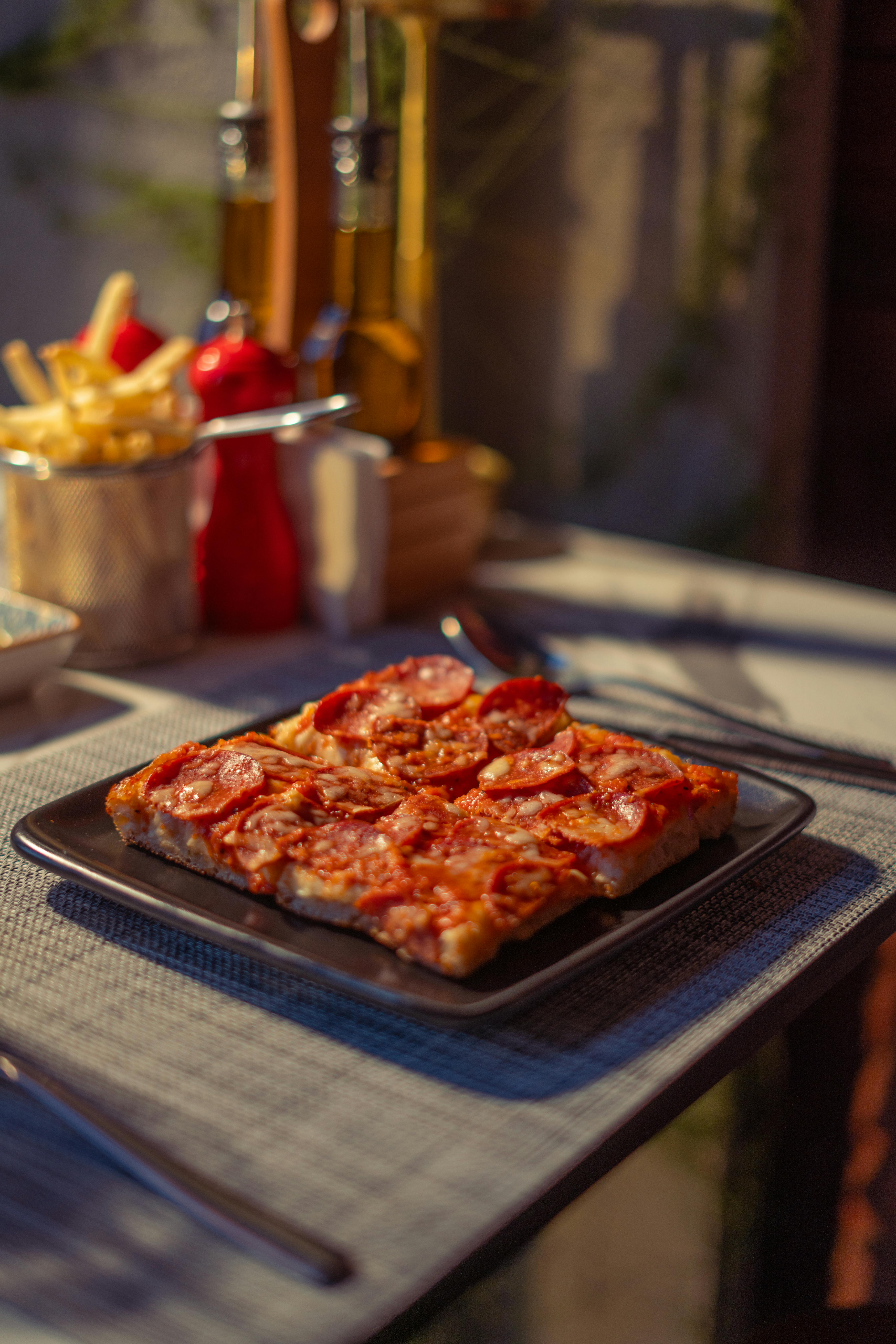 Square pepperoni pizza slice on table in cozy outdoor setting.
