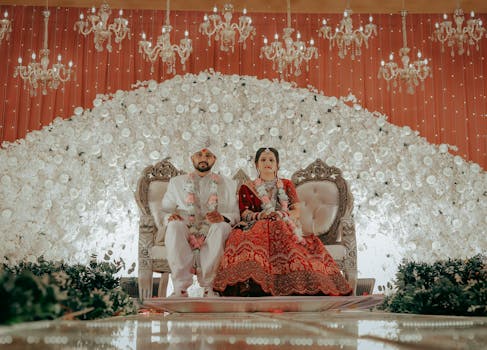 Beautiful Indian couple in traditional wedding attire seated on a decorated stage with floral backdrop.