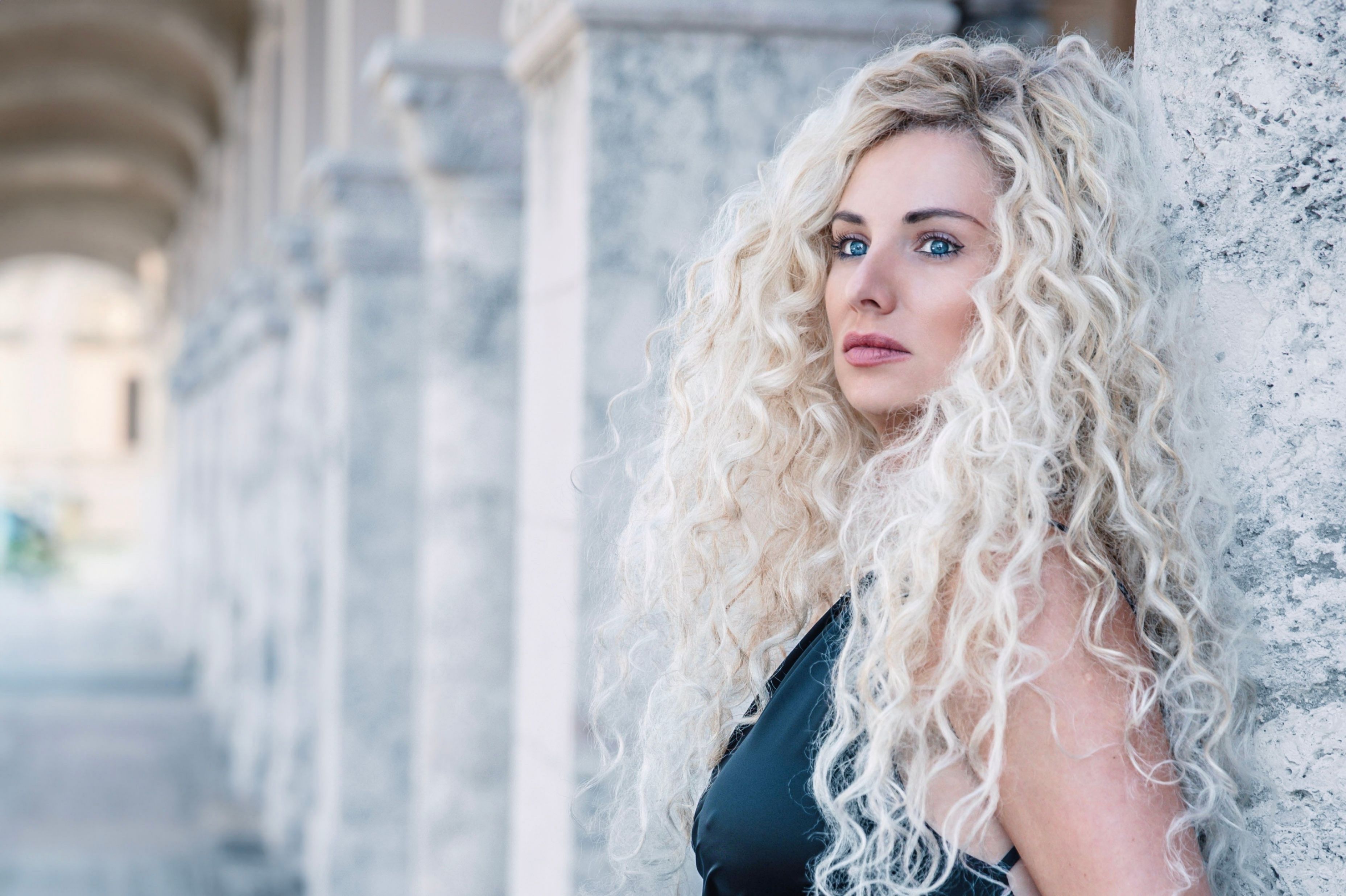 Stylish outdoor portrait of a woman with curly blonde hair leaning against a stone column. | BocaVibe