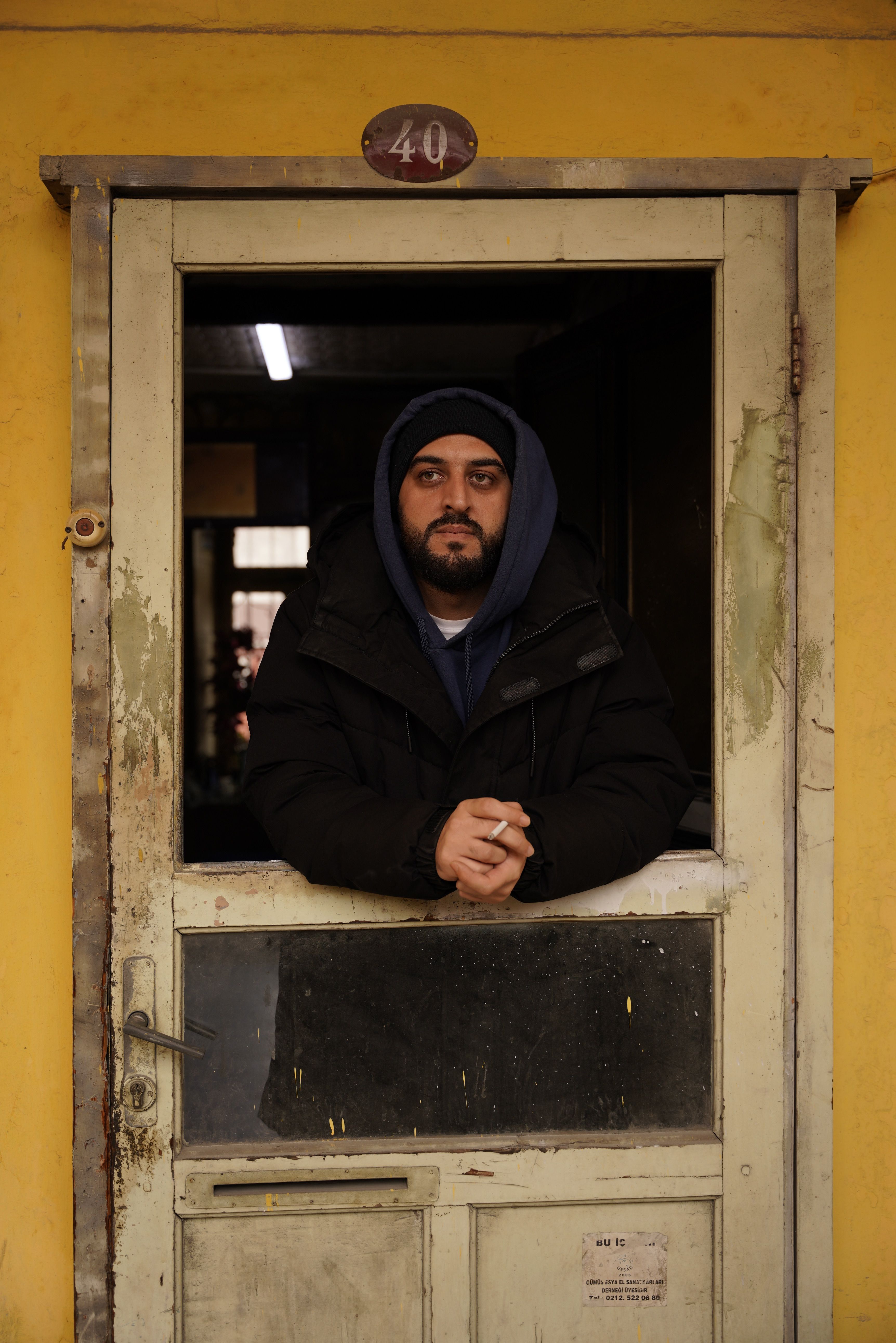 Man in a black coat and hoodie stands at a worn yellow door, looking contemplative.