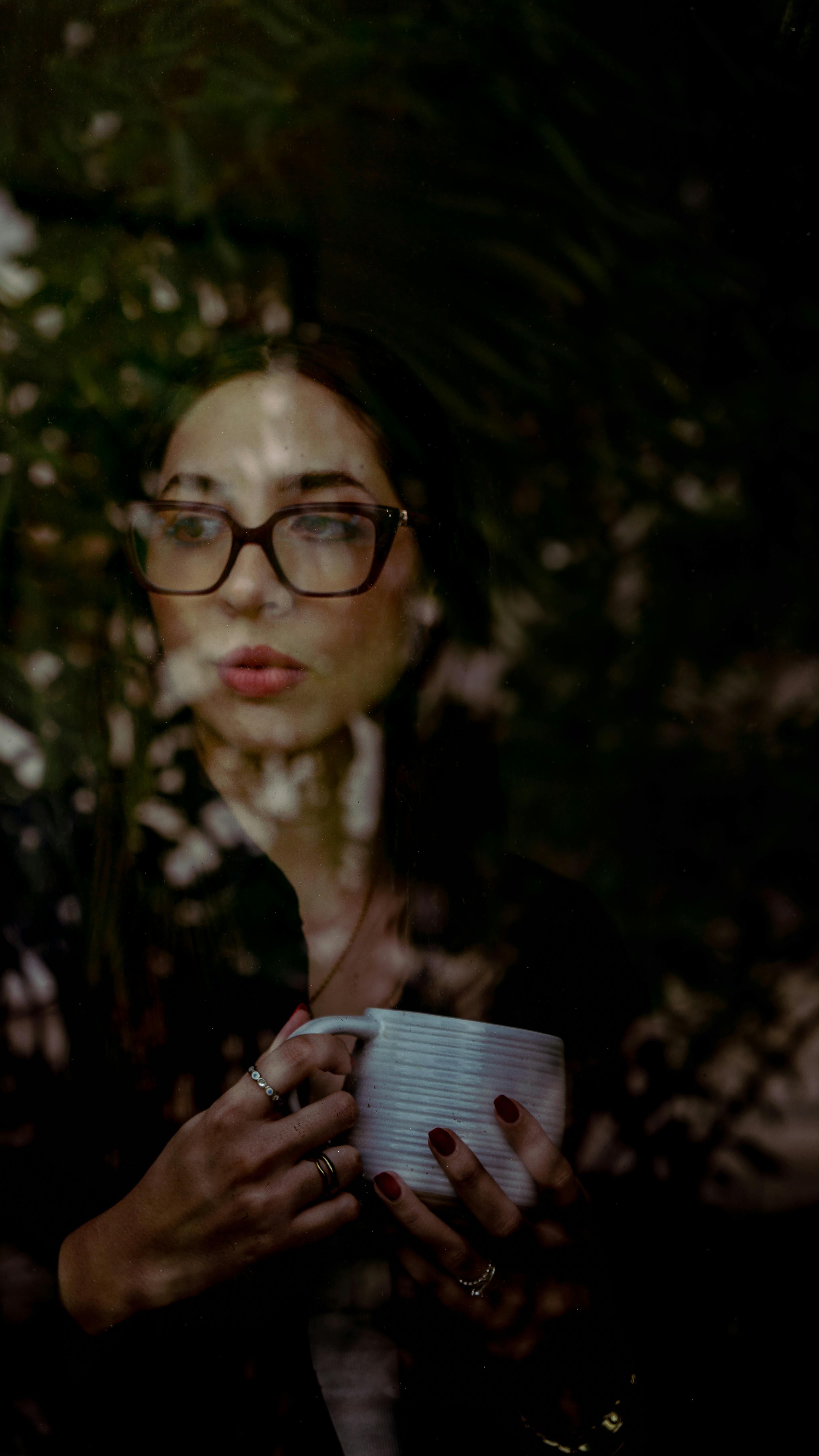 A pensive woman holding a coffee cup, seen through a window with reflections.