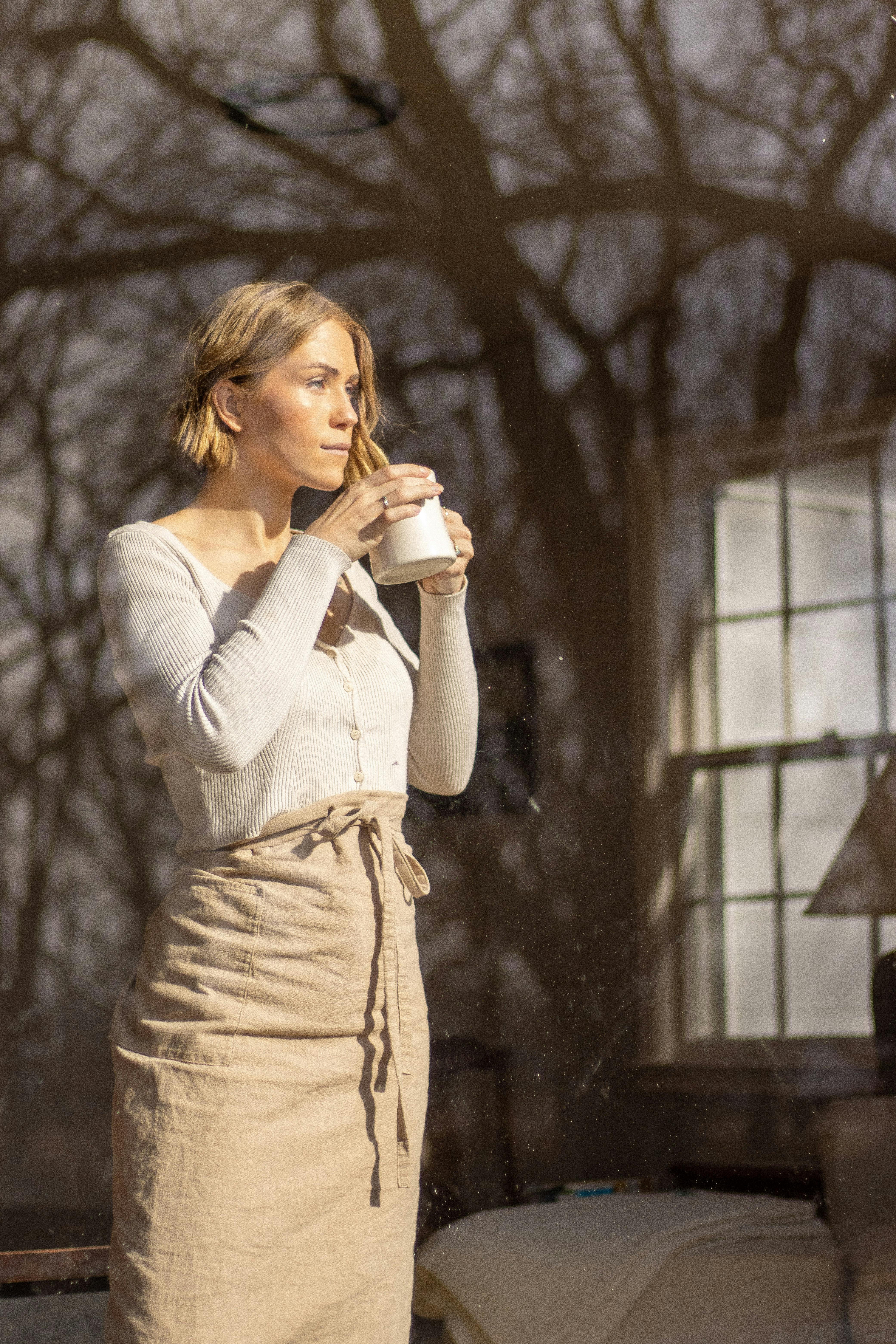 A blonde woman stands by a window, savoring a warm coffee, bathed in natural light.