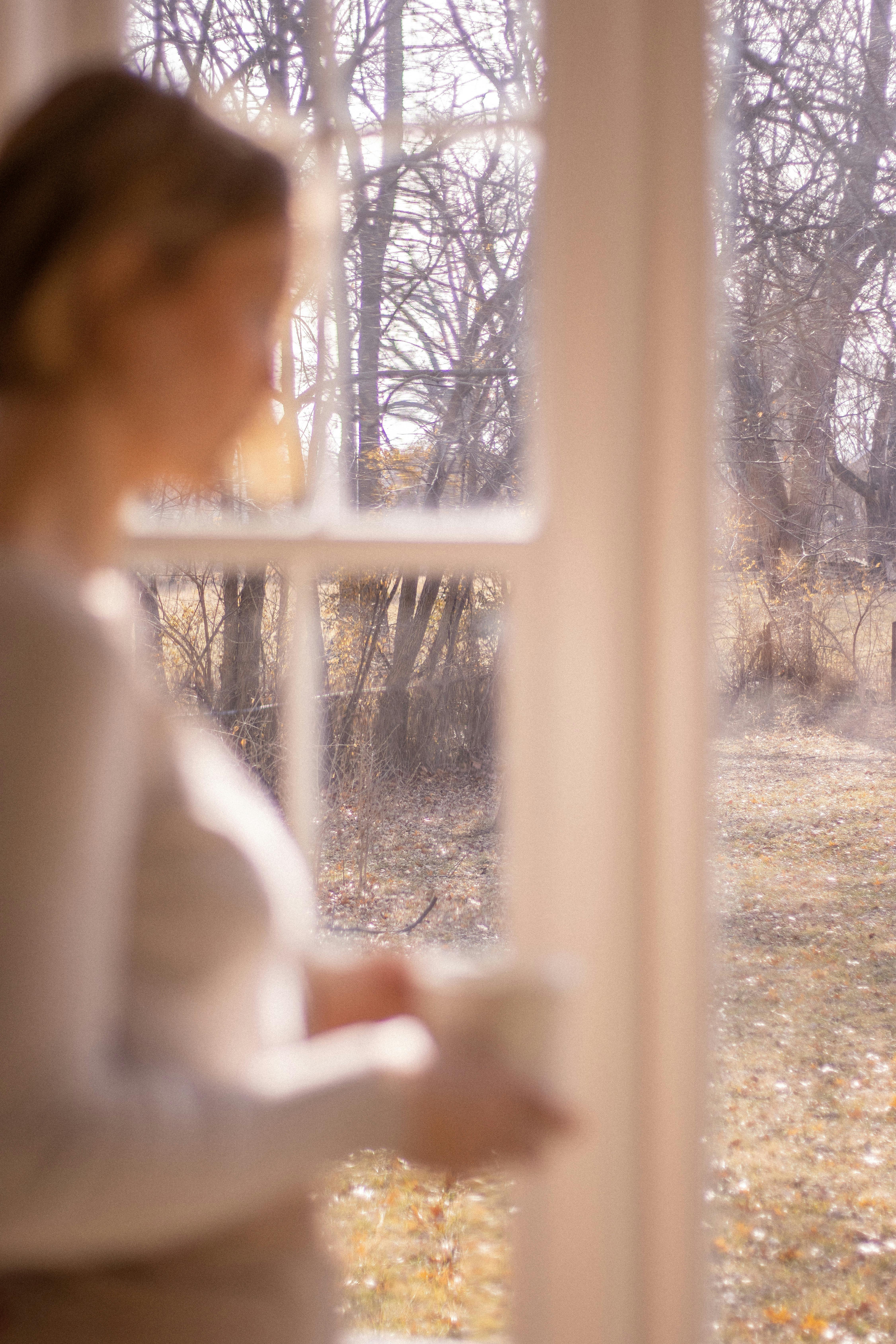 A woman stands by a window, looking outside at a sunlit autumn scene.