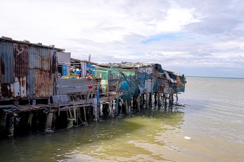 Free Rustic shanty homes on wooden stilts along a serene coastal shoreline with a partly cloudy sky. Stock Photo