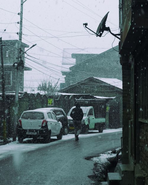 Free Winter street scene with falling snow, cars, and pedestrian, capturing serene urban atmosphere. Stock Photo