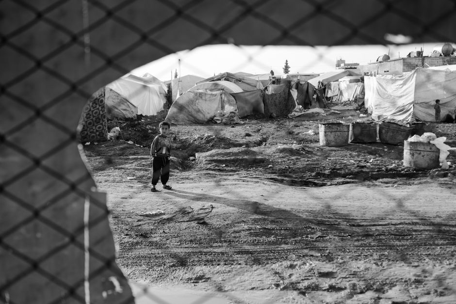 재난지원금 지급대상: 올해 내 이름도 포함됐을까? 2 A child stands alone in a Syrian refugee camp surrounded by tents, viewed through a fence.