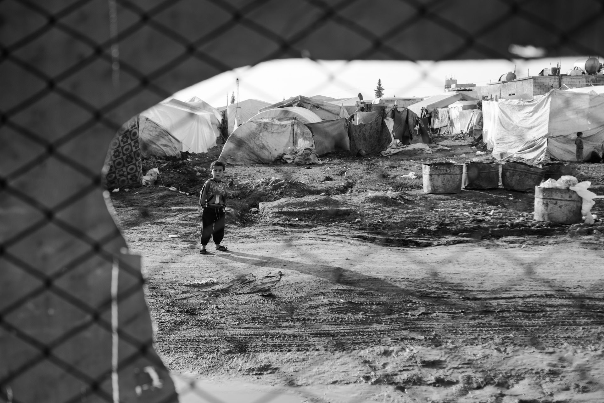 A child stands alone in a Syrian refugee camp surrounded by tents, viewed through a fence.