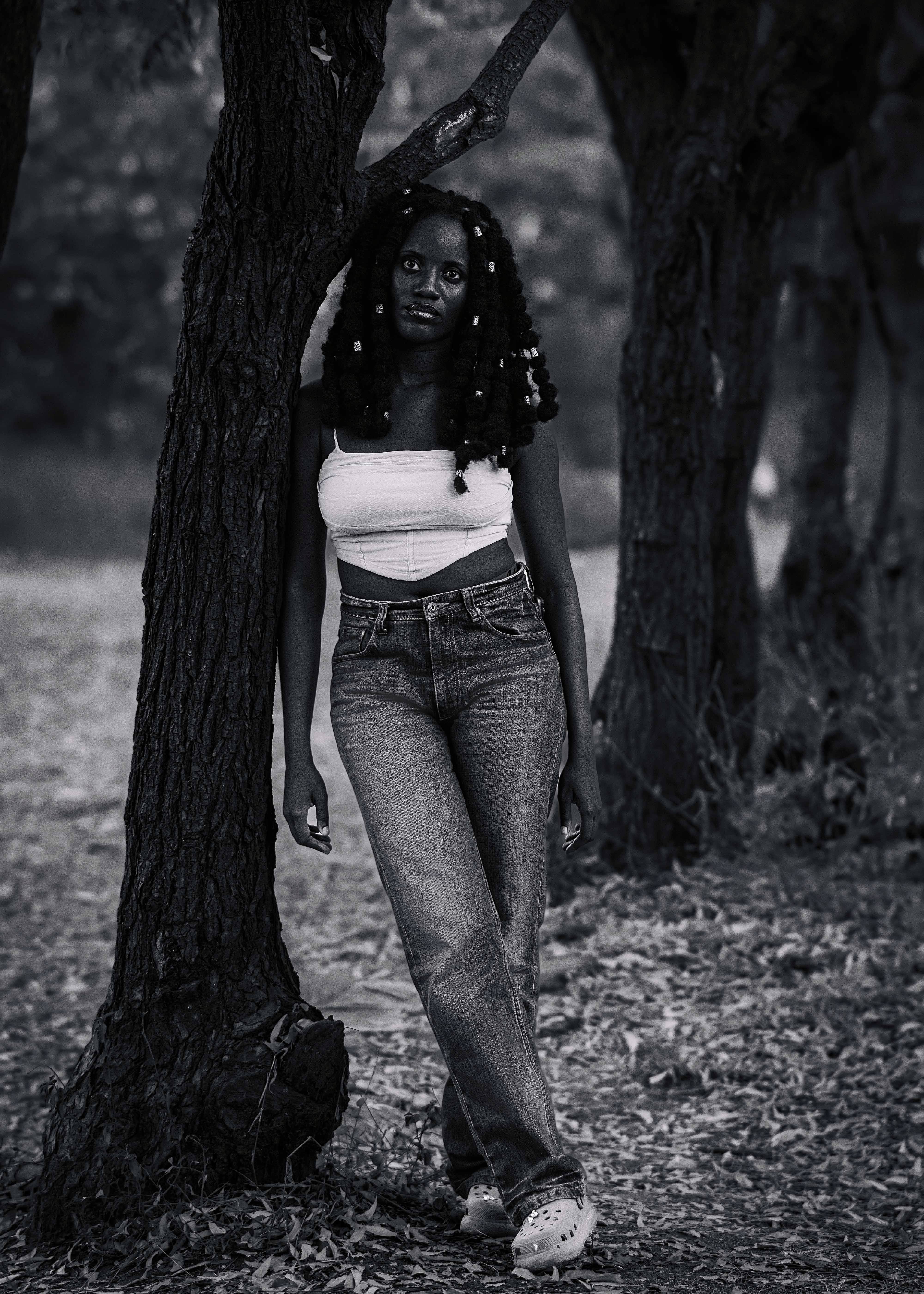 Black and white portrait of a woman leaning against a tree in a tranquil forest setting.