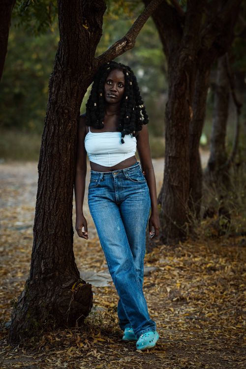 Free A young woman wearing casual attire leans against a tree in a serene park setting. Stock Photo
