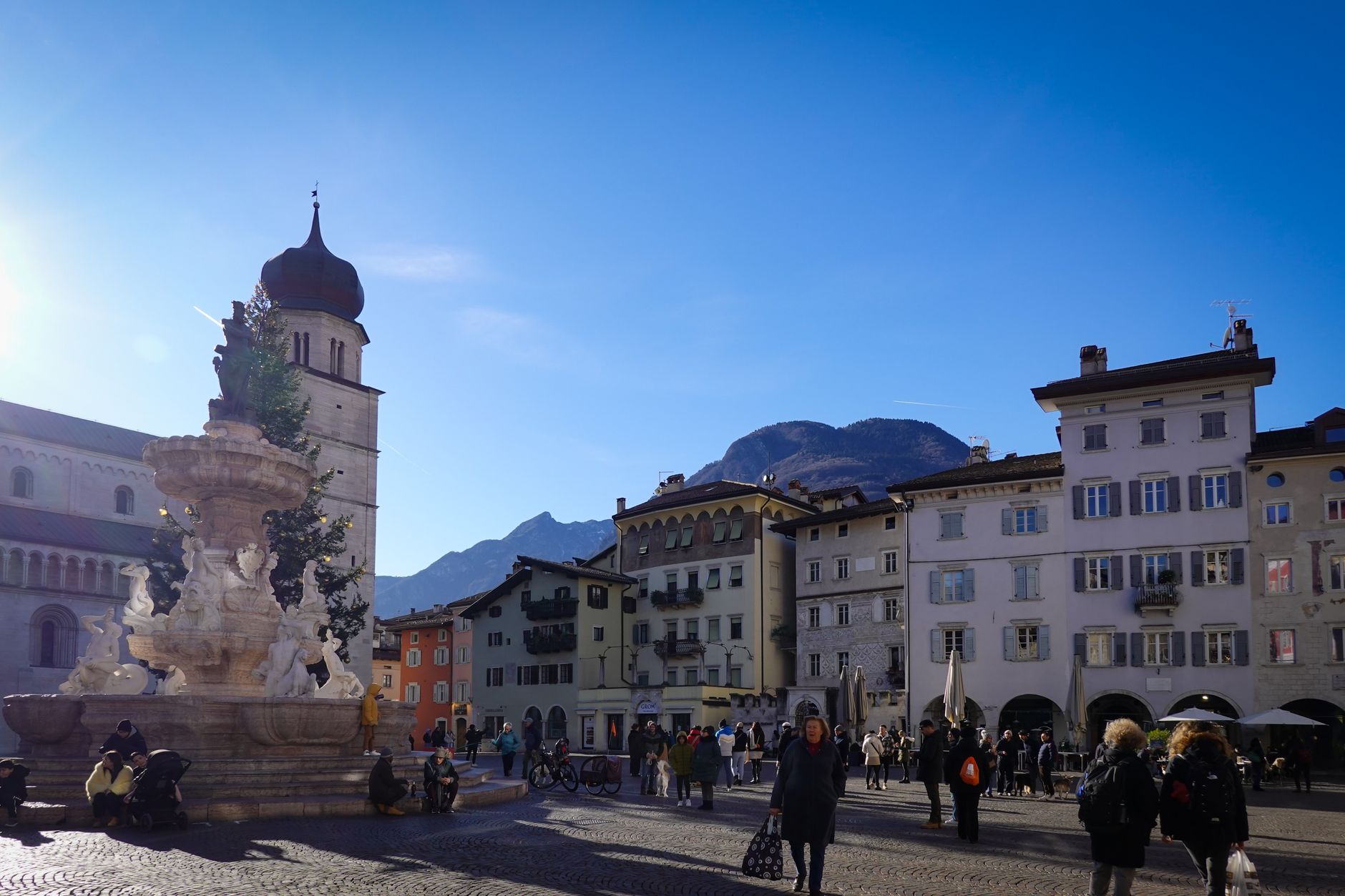 Charming Italian plaza with fountain and mountain backdrop, vibrant city life captured in natural light.
