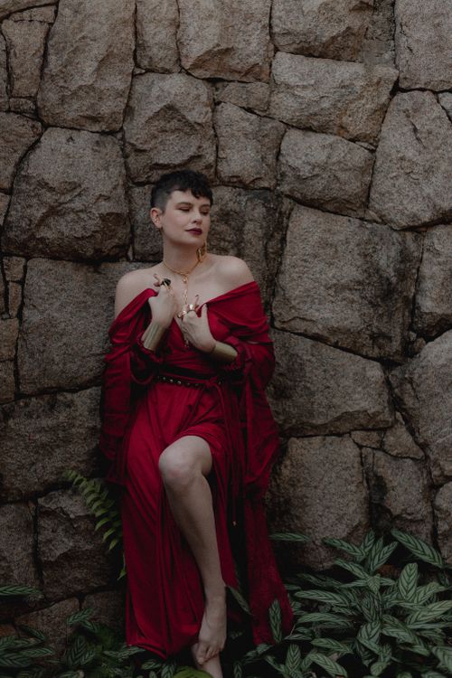 Free Woman in elegant red dress standing by a stone wall, looking serene. Stock Photo