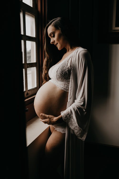 бесплатная Беременная женщина в кардигане у окна Стоковое фото