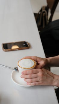 Hands holding a frothy coffee cup on a table with smartphone nearby, cozy setting.