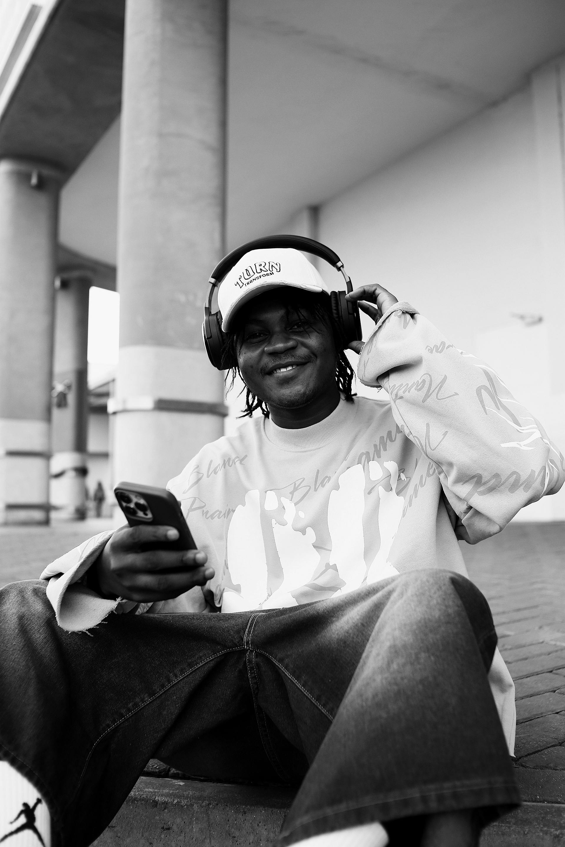 Black and white portrait of a man listening to music on headphones outdoors, smiling at camera.