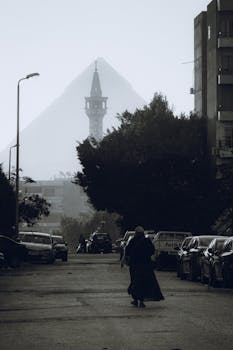 Black and white photo of a person walking in an urban street with a pyramid in the background.