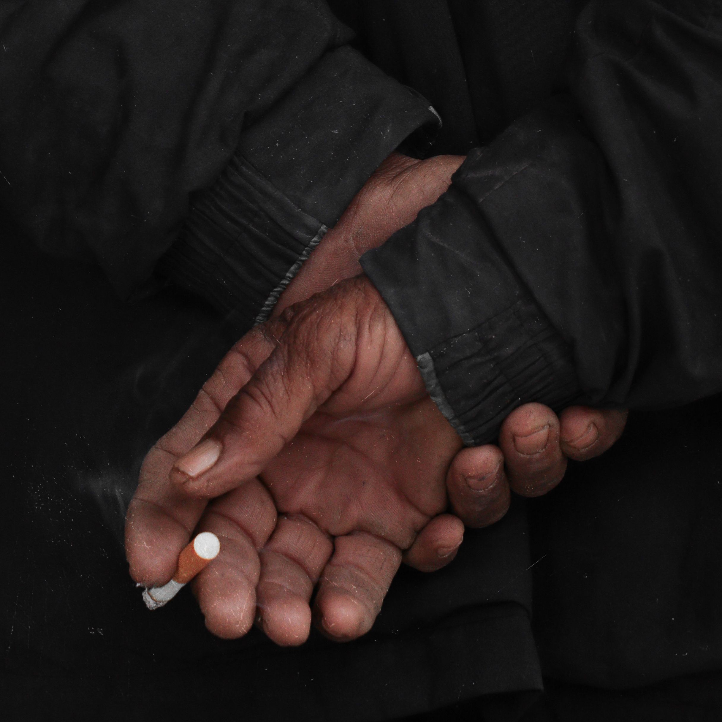 Close-up of older man's hands crossed, holding a cigarette, wearing a dark jacket.
