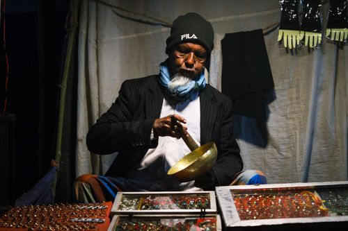 Free An elderly street vendor in Bangladesh plays a singing bowl, surrounded by colorful wares. Stock Photo