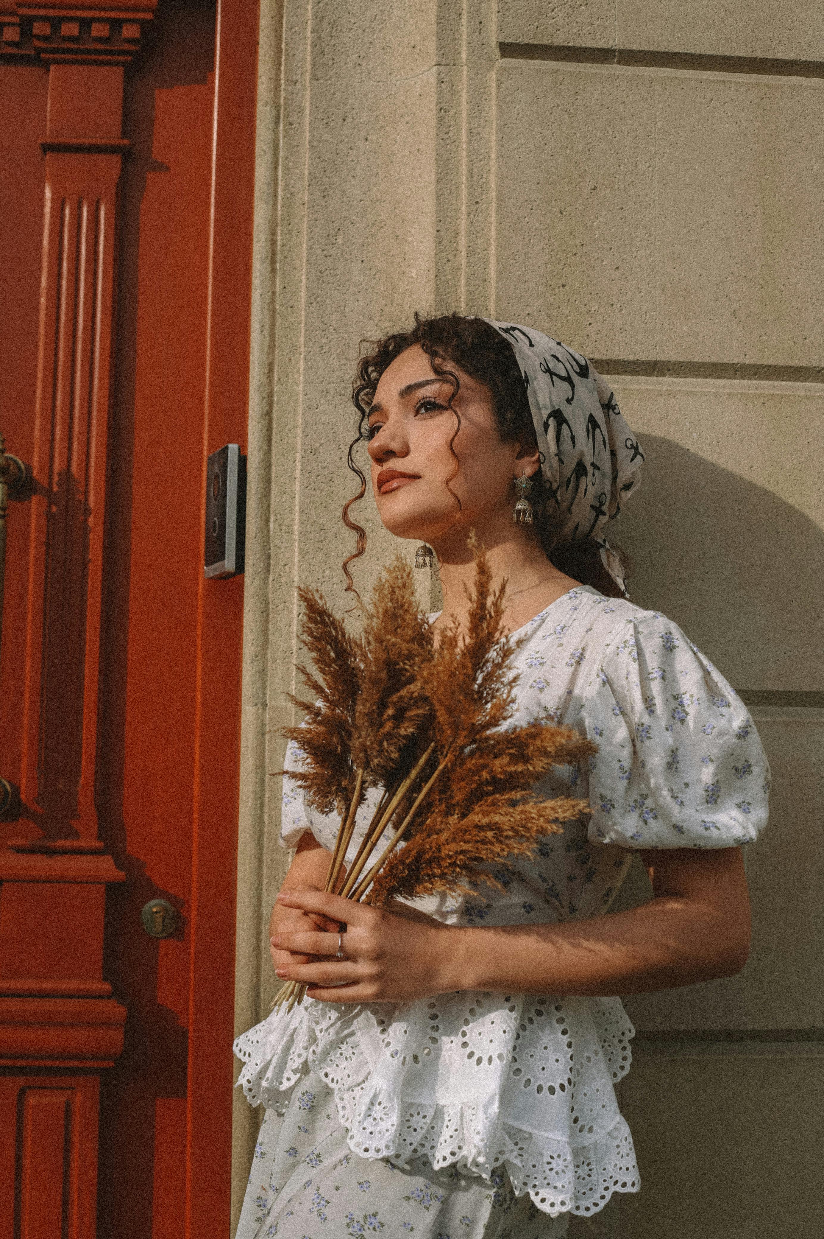 Elegant woman in vintage attire holding dry flowers by a red door, evoking a retro and artistic mood.