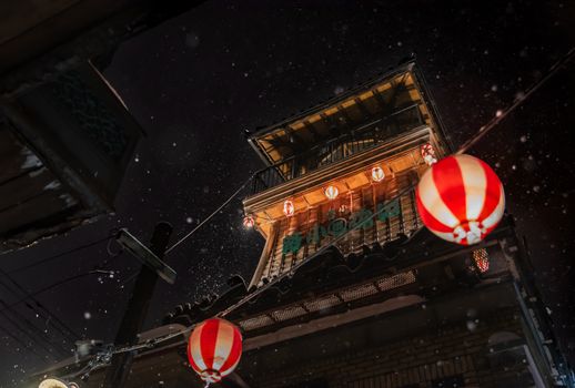 Illuminated traditional Japanese architecture with hanging paper lanterns during nighttime snowfall.