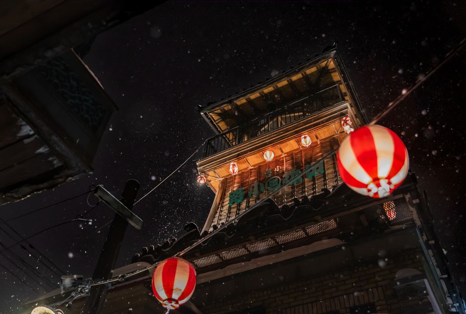 Illuminated traditional Japanese architecture with hanging paper lanterns during nighttime snowfall.
