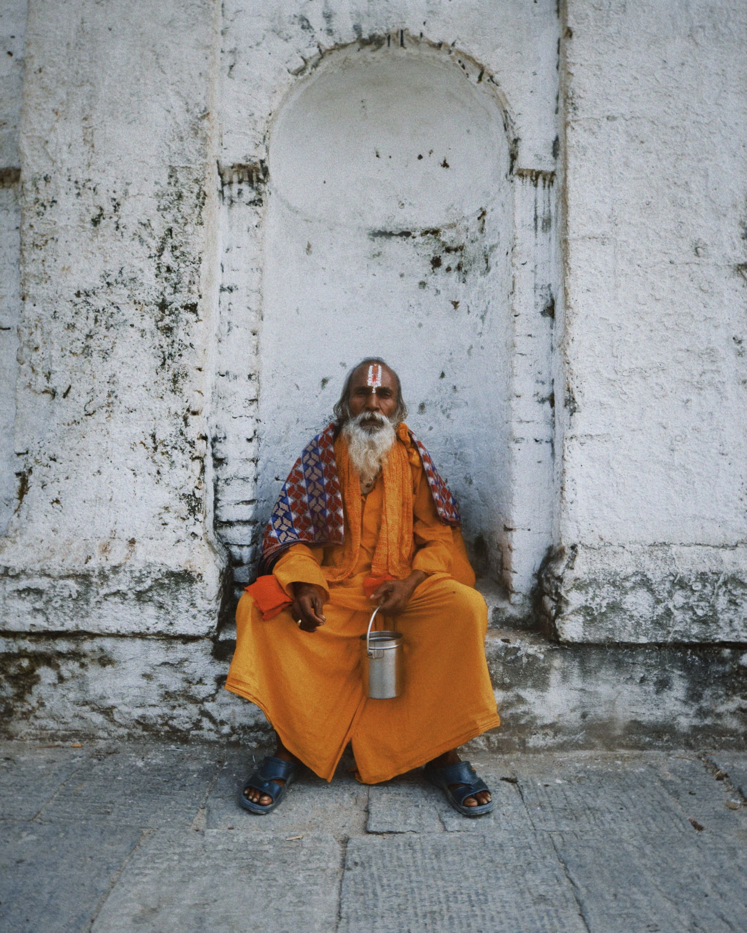 Elderly man in orange robe seated against white stone wall, holding metal container.