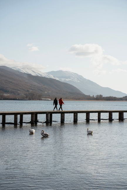 A tranquil scene of two people walking on a pier at Lake Ohrid, North Macedonia, in winter with swans in the foreground.