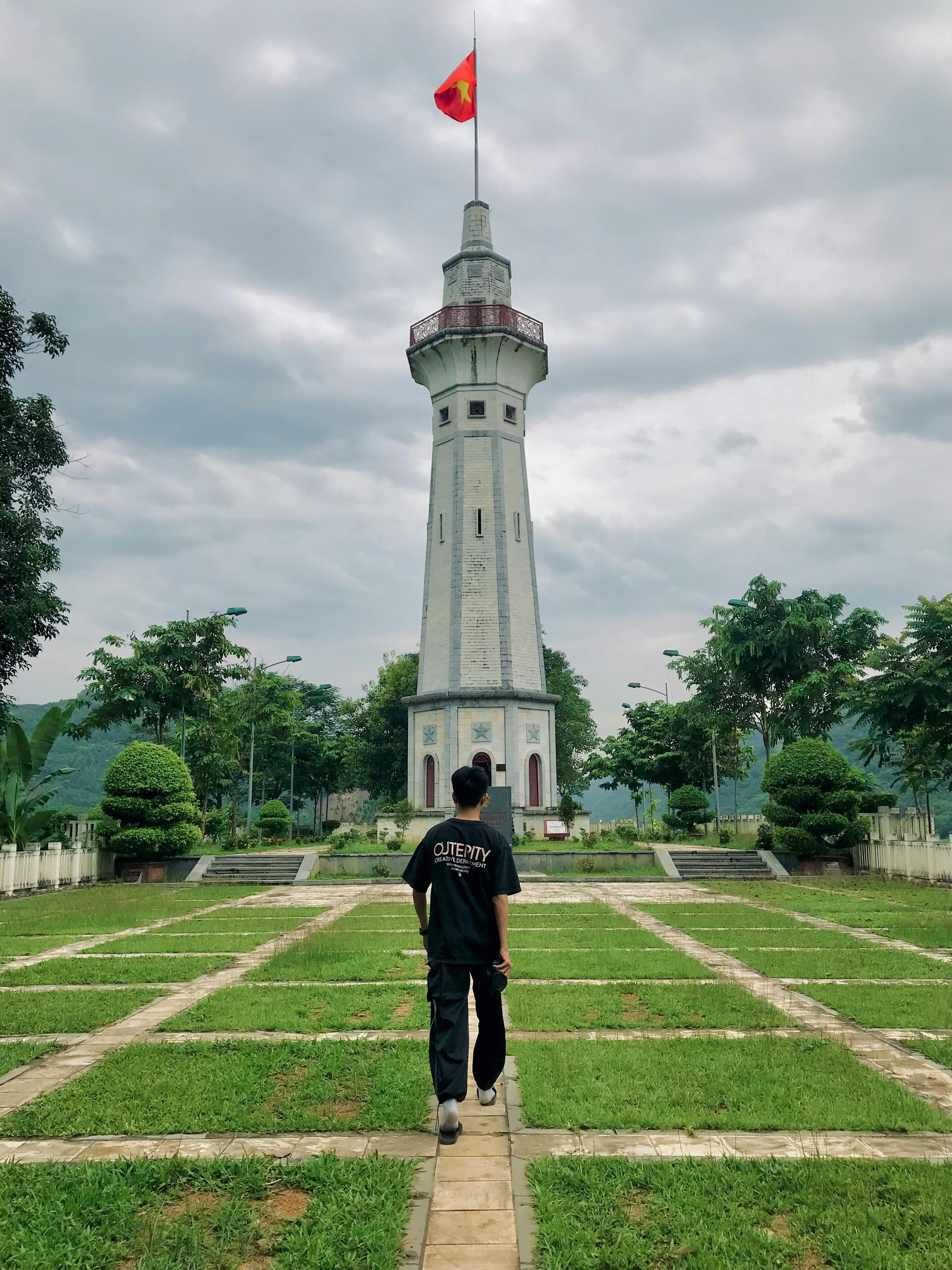 A person walking towards a tall tower with a red flag, surrounded by trees and cloudy sky.