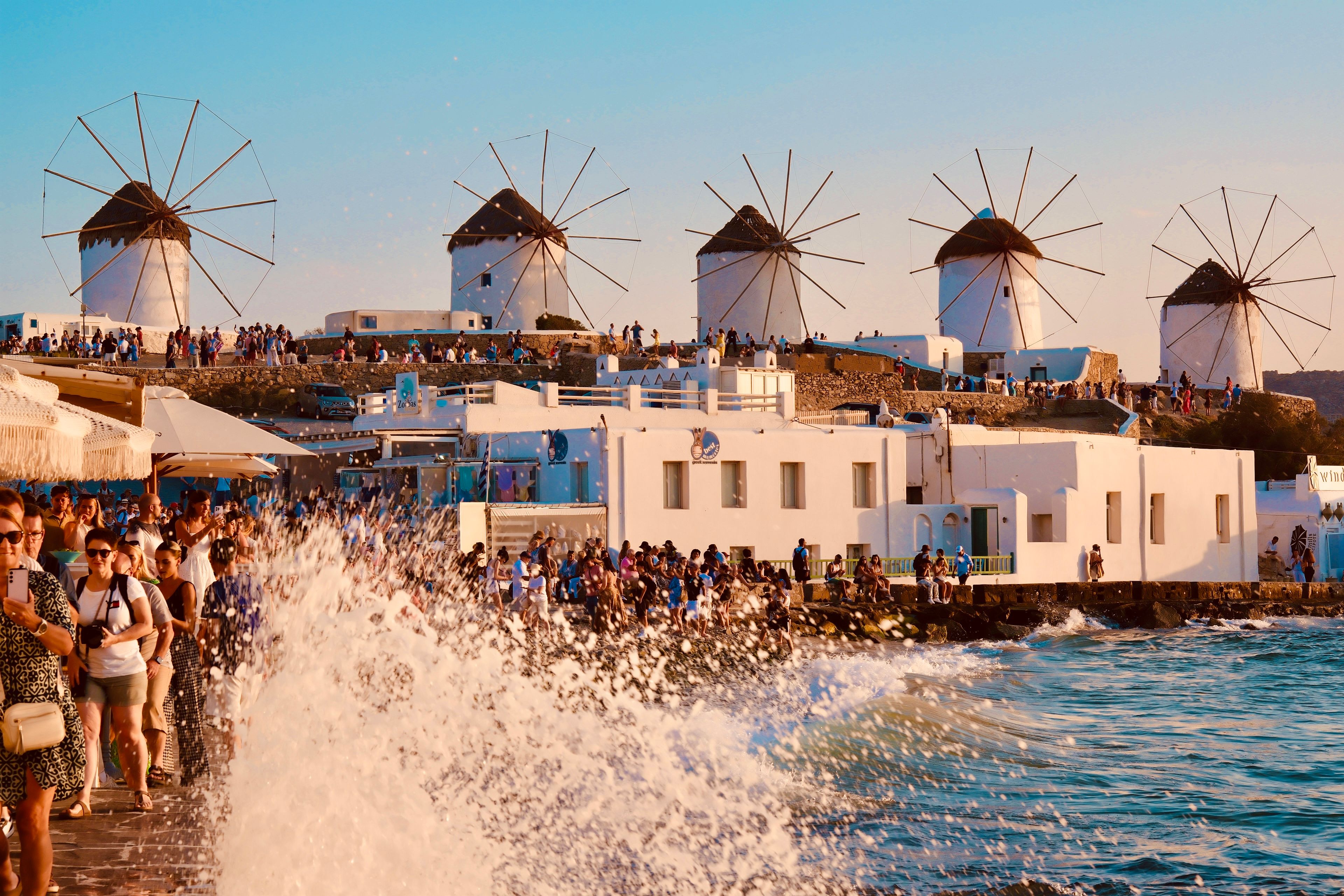 White windmills on a Mykonos hillside with crowds of tourists as waves crash along the seaside promenade at sunset