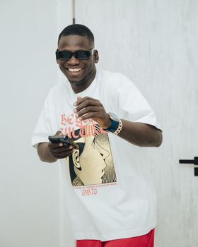 Cheerful man wearing sunglasses and stylish t-shirt, holding a smartphone indoors. - Fotografer Sidoarjo