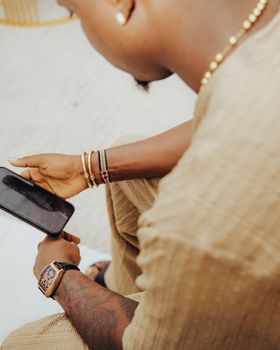 A young adult seated outdoors, adorned with trendy bracelets and a watch, looking at a smartphone.